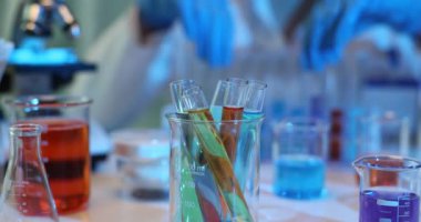 Renkli sıvılarla dolu çeşitli laboratuvar vitrinleri olan steril bir işyeri. Koruyucu eldivenli işçiler test tüplerini yavaş çekimde kontrol ediyor