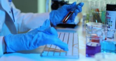 Bilim adamı bilgisayar üzerinde çalışıyor ve kırmızı sıvıyla tüpü tutuyor. Sıvıların laboratuvar testi