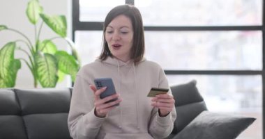 Evde kredi kartı ve akıllı telefon kullanan kendine güvenen bir kadın çevrimiçi alışveriş yapıyor. Çevrimiçi ödemeler ve alımlar çevrimiçi mağazalarda