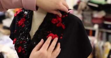 Terzi ellerinin, siyah pullu elbiseye kırmızı çiçek şeklinde pullar diktiği yakın plan. Terzi tasarımcısı manken üzerine yeni bir siyah elbise tasarlıyor.