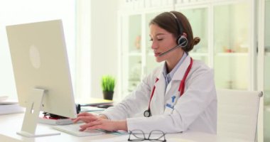 Bilgisayarda ağır çekimde kulaklık takan kadın doktor tipleri. Teletıp danışmanlığı yapan kadın hasta notlarını kopyalıyor.