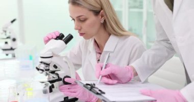 Kadın asistan, tıp laboratuarında mikroskoba bakan bilim adamına kayıtları gösteriyor. Meslektaşlar laboratuardaki iş arkadaşlarıyla test sonuçlarını kontrol eder
