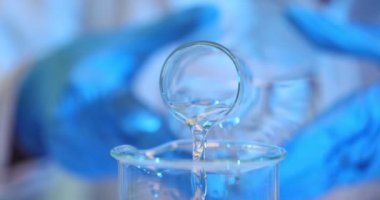 Koruyucu eldivenli dikkatli bilim adamı şeffaf beher kabına yavaş çekimde su döküyor. Yetenekli işçiler deney için bileşik hazırlar.