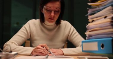 Yorgun kadın, akşam ağır çekimde ofiste çalışırken hayati önem taşıyan bir rapor hazırlıyor. Yorgun kadın işçi kafasını klasör yığınıyla masaya koyuyor.