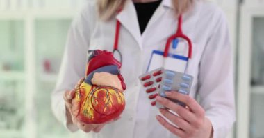 Kadın doktor kalp modelini tutuyor ve klinikte haplar ağır çekimde su topluyor. Kardiyolog, yenilikçi ilaçların hastalara sağladığı faydaları anlatıyor