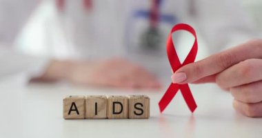 Doktor küplerin yanında kırmızı kurdele tutuyor ve masada AIDS yazıyor. Kadın, salgın hastalıkla mücadele ve destekle mücadelenin devam ettiğini hatırlatıyor.