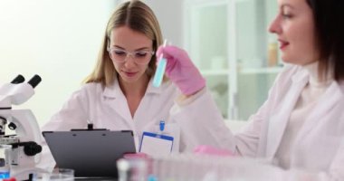 Kadın laboratuardaki meslektaşına renk sıvısıyla test tüpü gösteriyor. Kadın kimyagerler beklenmedik bir sıvı reaksiyonuna şaşırdılar. Tıbbi araştırma