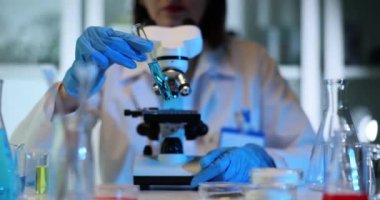 Kadın tıp çalışanı laboratuarda mavi reaktörle test tüpünü sallıyor. Kadın bilim adamı, kimya laboratuvarında ağır çekimde renk sıvısı malzemesi karıştırıyor.