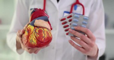 Doktorun elinde ilaç tabletleri ve yapay kalp modeli var. Doktor kalp sağlığı için yavaş çekimde ilaç almanın önemini gösteriyor.