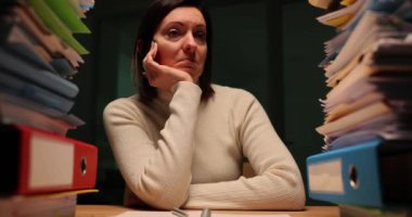 Kadın ofis çalışanı, akşamları iş yerinde duran raporlarla dolu dosyalara bakıyor. Yorgun kadın görev hacmiyle sinir ediyor.