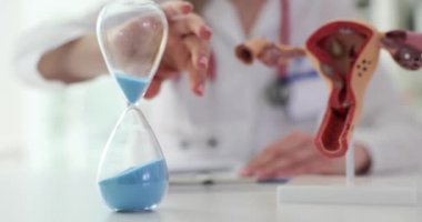 Kadın doktor, klinikte rahim modelinin yanında kum saati kullanıyor. Jinekolog eski usul saatle hasta bakımını optimize ediyor.
