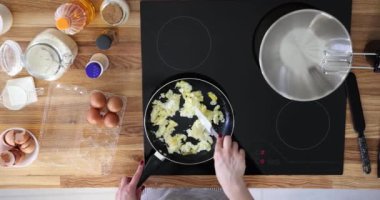 Önlüklü bir kadın, ağır çekimde spatula kullanarak yumurta pişirir. Kadın modern mutfakta indüksiyon sobasıyla yemek pişirmeye başlıyor.