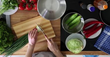 Önlüklü bir kadın yavaş çekimde yeşil soğan doğruyor. Kadın modern mutfakta taze salata için zengin bir malzeme hazırlıyor.