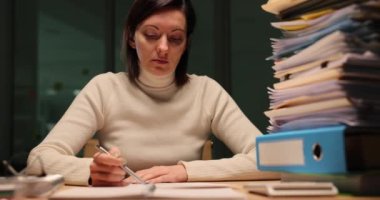 Ofisteki sayısız belgeyi incelerken çalışmaktan ve not almaktan yorgun düşmüş bir kadın yönetici. Fazla mesai yorgunluğu ve teslim tarihi