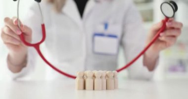 Doktor, klinikteki hastaları temsil eden ahşap heykelcikleri incelemek için steteskop kullanıyor. Sağlık sigortası ve kalp hastalığı tedavisi