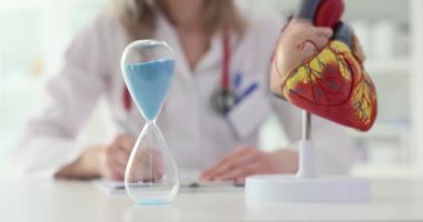 Kalp tedavisinin anatomik modelini inceleyen ve kalp tedavisinin önemini simgeleyen kum saatini gözlemleyen kardiyolog. Kalp hastalığı belirtileri ve tedavi süresi