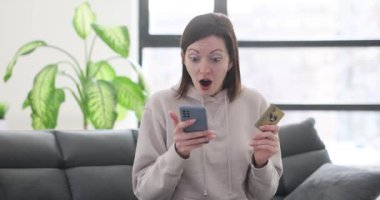 Oturma odasında elinde kredi kartıyla telefon ekranına bakan bir kadın. Dolandırıcılar mobil bankacılık yoluyla para çalıyorlar.