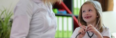 Meraklı küçük kız steteskopu doktorlara yardım etmek için hazır tutuyor. Uzman, çocuğa klinikte sağlık ekipmanı kullanmayı öğretiyor.