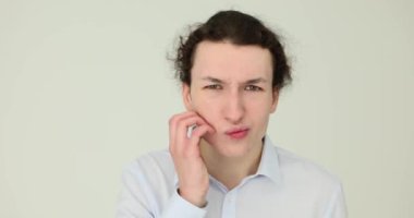 Yüzünde somurtkan bir ifadeyle beyaz arka planda kaşınan genç bir adam. Yüz alerjisi ve kızarıklıkları.