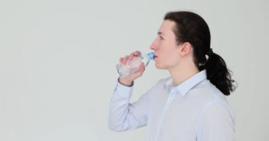 Genç bir adam beyaz arka planda plastik bir şişeden su içiyor. İçki rejimi ve susuzluğun önlenmesi
