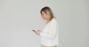 Beyaz arka planda telefonla mesajlaşan genç bir kadın. Akıllı telefon tutan ve gergin bir şekilde sosyal medyada gezinen kadın.