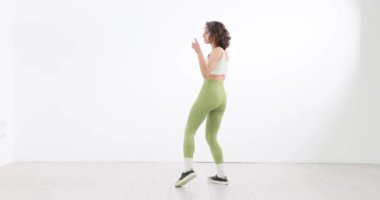 Stüdyoda yeşil taytlarla dans eden kadın beyaz arka planda. Kız kendine güveni ve coşkusu ile hareket eder.