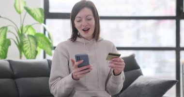 Mutlu kadın oturma odasında kanepede otururken online alışveriş yapmak için akıllı telefon kullanıyor. Elinde banka kartı ve telefon tutan gülümseyen kız.