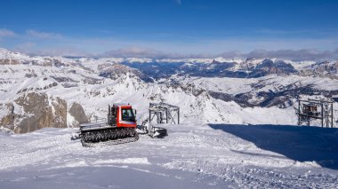 Bu resim, açık mavi bir gökyüzünün altındaki kar kaplı bir dağ sırasının nefes kesici bir görüntüsünü gösteriyor. Karlı araziler için tasarlanmış kırmızı bir kar aracı ön planda belirgin bir şekilde yer almaktadır..