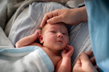 Mavi bir battaniyeye sarılmış bir bebek, uzanmış ve annesine yukarıdan bakıyor. Annenin eli bebeğin kafasına hafifçe dokunur, bir anlık bakım ve bağ kurar.. 