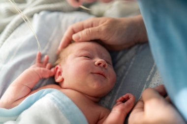 Mavi bir battaniyeye sarılmış bir bebek, uzanmış ve annesine yukarıdan bakıyor. Annenin eli bebeğin kafasına hafifçe dokunur, bir anlık bakım ve bağ kurar.. 