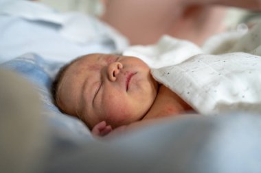 Beyaz bir battaniyeye sarılı, huzur içinde uyuyan yeni doğmuş bir bebeğin huzurlu bir görüntüsü. Sakin ve sessiz ortam, bebeklerin huzur ve güvenliğinin altını çiziyor..