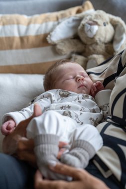 Yeni doğmuş bir bebek aile fertlerinden birinin kollarında bir yatakta, arkasında oyuncak bir ayıyla dinleniyor. Görüntü, bir aile ortamının sıcaklığını ve rahatlığını ön plana çıkarıyor. Bebek yorgun ya da uykuda görünüyor..