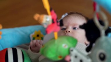 Canlı bir bebek yumuşak, renkli bir minder üzerinde oynuyor, merak ve bağlılık sergiliyor. Açık kahverengi saçlı ve büyük mavi gözlü bebek, canlı oyuncaklarla çevrili..