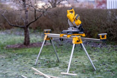 Bu görüntü, arka planda çimen ve çalıların bulunduğu sakin bir ortamda sağlam bir standın üzerine monte edilmiş sarı bir elektrikli testereyi gösteriyor..