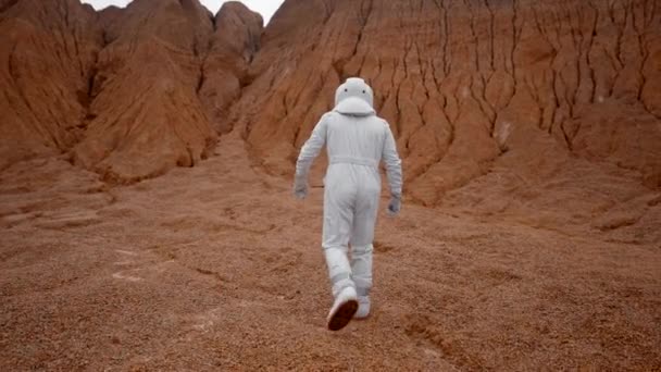 白いスーツを着た宇宙飛行士の後ろからの眺めと赤い丘に向かって歩くヘルメット 未知の惑星のクレーターの中の宇宙飛行士は表面を探検しに行きます — ストック動画