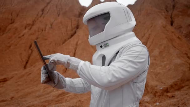 宇宙飛行士は遠くの赤い惑星の表面でタブレットで作業しています 白いヘルメットとデジタルパッドで作業スーツの白人男性 将来の植民地化の新しい地平線を探索 — ストック動画