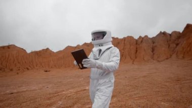 Mars 'ın yüzeyinde elinde bir tabletle bir astronot yürüyor. Koruyucu beyaz giysi ve kask atmosferin yokluğunda nefes almasına yardımcı oluyor.