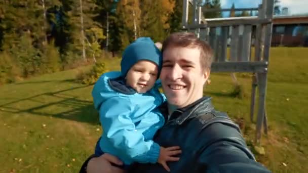 腕の中で息子を抱えカメラを見ている父親のハメ撮り 男Recordsビデオの回転とともに幼児でスマートフォン上の公園遊び場クローズアップ — ストック動画