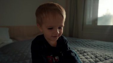 Meraklı küçük çocuk, kitaptaki resimlere işaret ediyor. Parlak pencereye karşı yatakta oturuyor. Küçük çocuk sabahını yatak odasında yavaş çekimde geçirir.