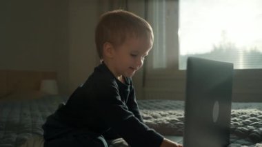 Tatlı çocuk modern dizüstü bilgisayarın klavyesinde yazı yazmayı öğreniyor. Meraklı çocuk, yatak odasındaki aleti yavaş çekimde kullanmada yeni yetenekler kazanıyor.