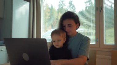 Önemseyen anne, kucağında oğluyla modern bir dizüstü bilgisayarda çalışıyor. Odaklanmış kadın serbest çalışan, mutfak masasında oturan cihaza bakar.