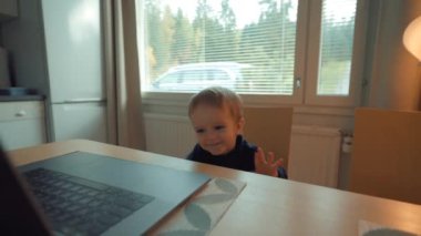 Sevimli küçük çocuk modern dizüstü bilgisayarını kullanmak için mutfak masasında oturuyor. Sarışın çocuk evde yavaş çekimde elektronik cihaza ilgi gösteriyor.