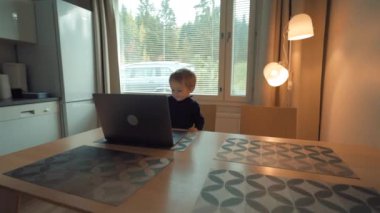 Tatlı çocuk parlak pencereye karşı açık dizüstü bilgisayarın yanında şık bir mutfak masasında oturuyor. Küçük çocuk ağır çekimde modern aletlere ilgi gösteriyor.