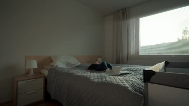 少年は足で本を蹴って柔らかいベッドの上に横たわっている ケアフリー幼児は明るい窓のあるモダンなコテージハウスのベッドルームにあります — ストック動画