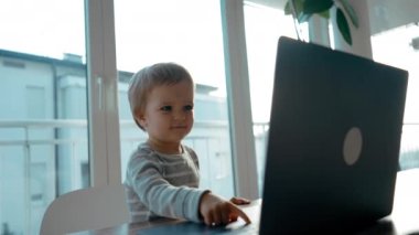 Meraklı küçük çocuk panoramik pencereye karşı masada oturan modern laptop ekranına bakıyor. Şirin çocuk elektronik cihaz kullanıyor.