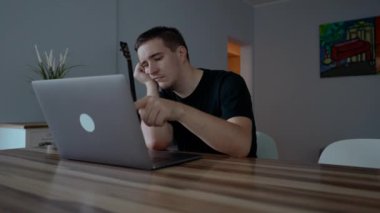 Yorgun adam modern dizüstü bilgisayarın tuşlarına basıyor. Genç serbest çalışan, oturma odasındaki ahşap masada otururken tembellik ediyor.