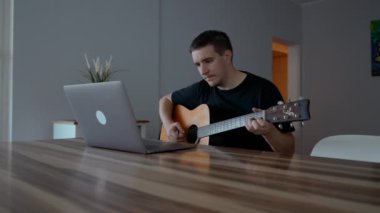 Adam dizüstü bilgisayarda eğitim videoları izlerken gitar çalıyor. Acemi müzisyen ahşap masada ağır çekimde müzik aleti kullanmayı öğreniyor.