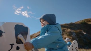 Mutlu çocuk Highland 'da büyük sallanan köpeğe binmeyi seviyor. Tatlı küçük çocuk güneşli bir günde İsviçre dağlarına giderken eğleniyor.