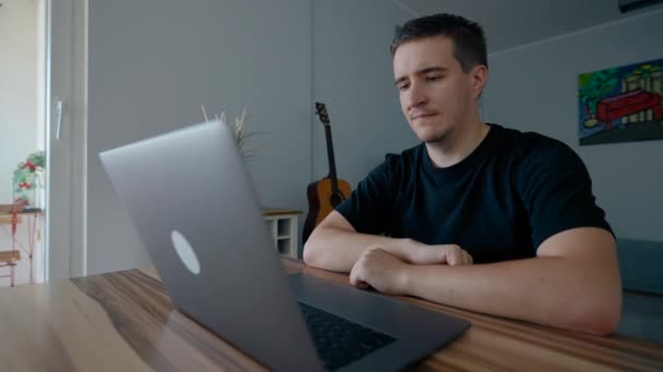 焦点を当てた男は木製のテーブルに座ってノートパソコンを介して興味深い講義を聞きます 男性学生は自宅で電子機器を介してオンライン教育を受ける — ストック動画
