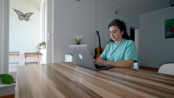 ブルネットの女性は現代のラップトップに木製のテーブルタイプの記事に座っています 若いフリーランサーは 電子機器のスローモーションを介して自宅から働くことを楽しむ — ストック動画